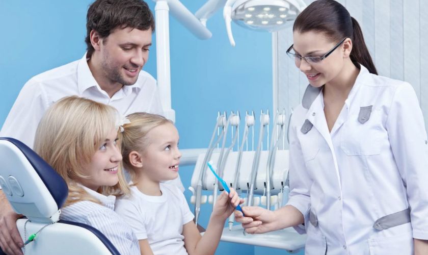 Dentist showing dental tools to young children and parents in modern dental office.