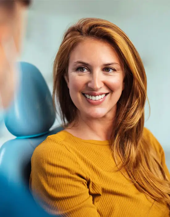Smiling woman with reddish-brown hair in yellow shirt next to blue dental chair.