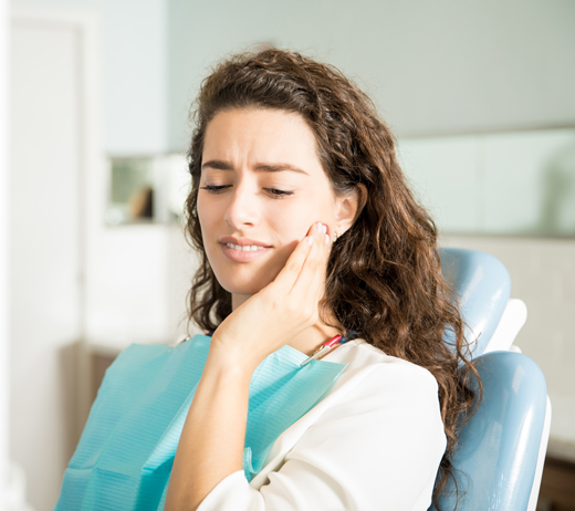 Woman with wavy brown hair in dental chair wearing bib, hand to face, smiling.