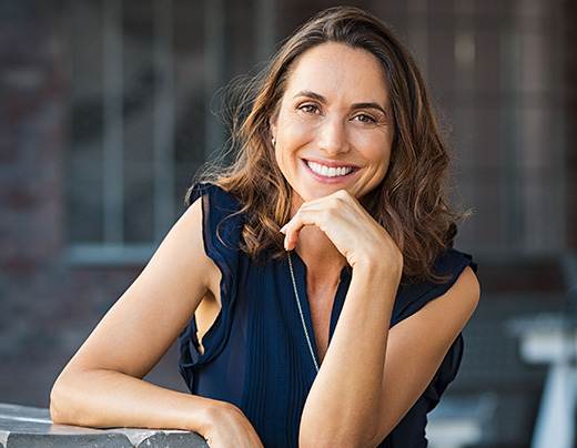Professional woman smiling at camera, resting chin on hand, wearing navy sleeveless top in urban setting.