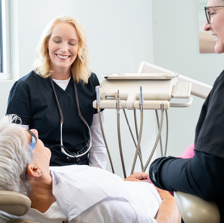 Dentist in black scrubs smiling while examining patient in dental chair with equipment visible.