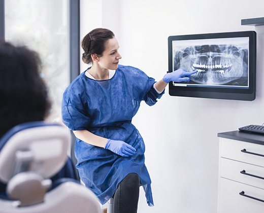 Dentist in blue scrubs explaining dental X-ray to patient in clinical office setting.
